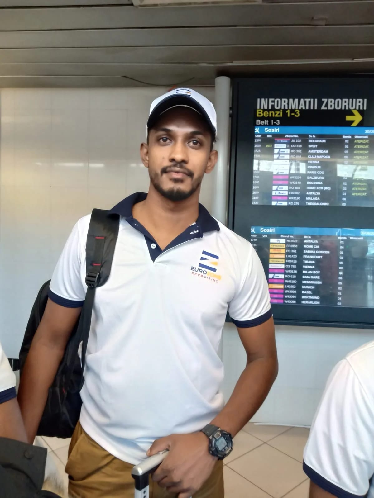 A man in a Eurozone polo shirt and cap, a backpack over one shoulder and a suitcase in hand, standing in front of a Romanian airport arrivals board