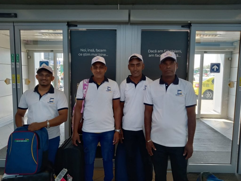 Four men in matching Eurozone polo shirts and caps standing with their luggage at the doors of a Romanian airport terminal