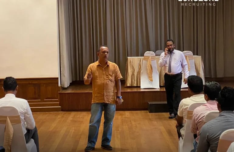 A colleague speaking to a seated group in a function room while another takes a call at the back.
