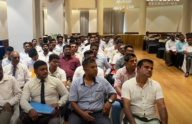 Rows of men in light shirts seated close together in a chandeliered function hall, facing the front, several holding document folders