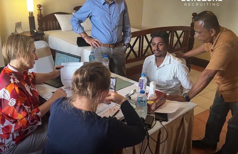 A candidate in a white shirt interviewed across a table in a hotel room, one interviewer reading his papers beside a laptop, another holding up a phone