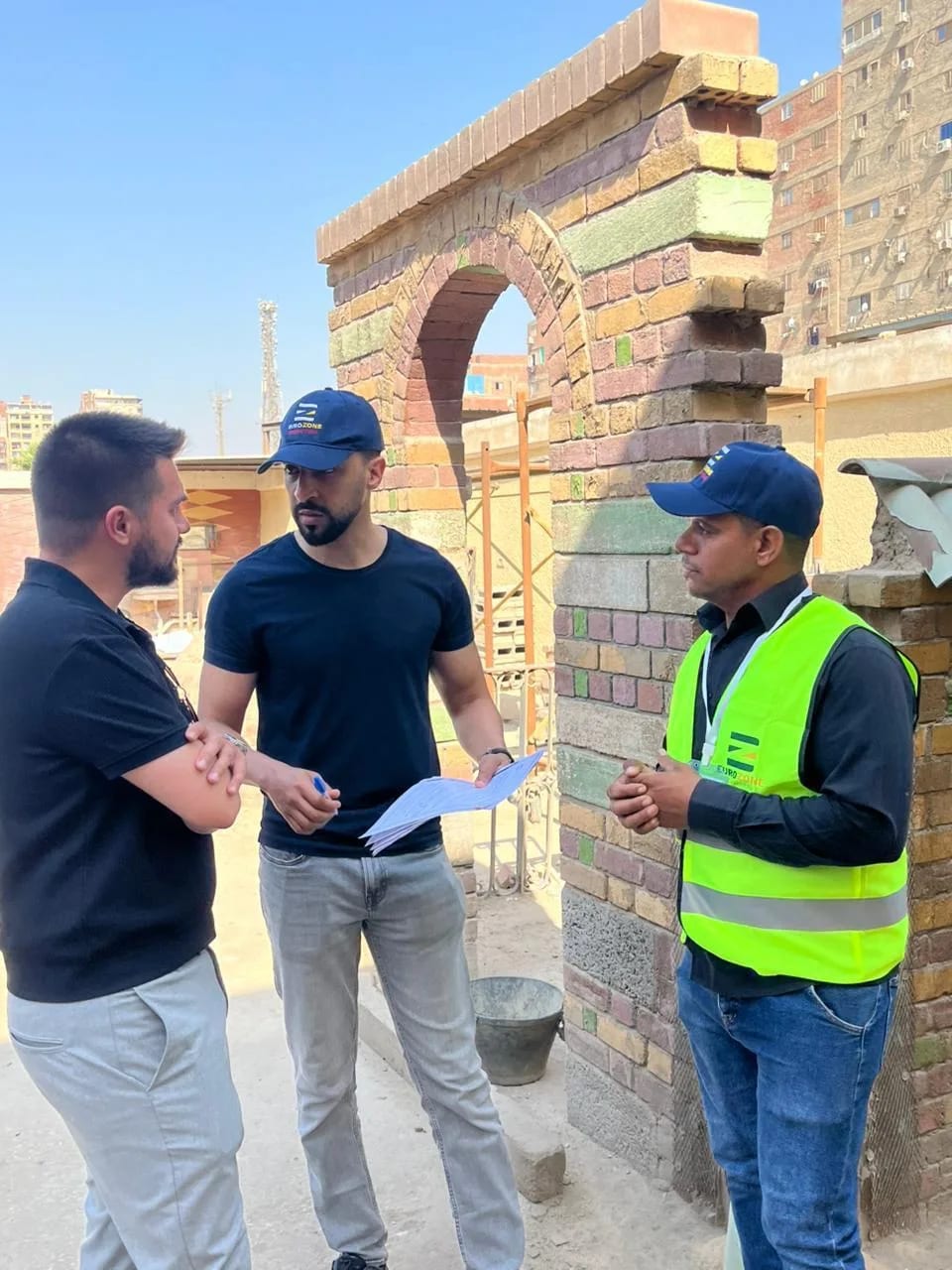 Trei bărbați discutând lângă o arcadă de cărămidă în construcție pe un șantier, unul ținând un teanc de documente, altul purtând o vestă reflectorizantă Eurozone