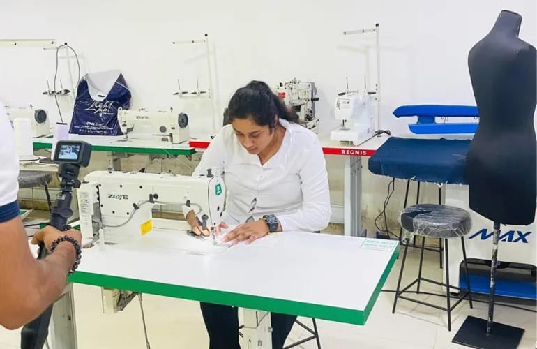 A woman guiding fabric under the needle of an industrial sewing machine, in a training workshop lined with more machines and a tailor's dummy